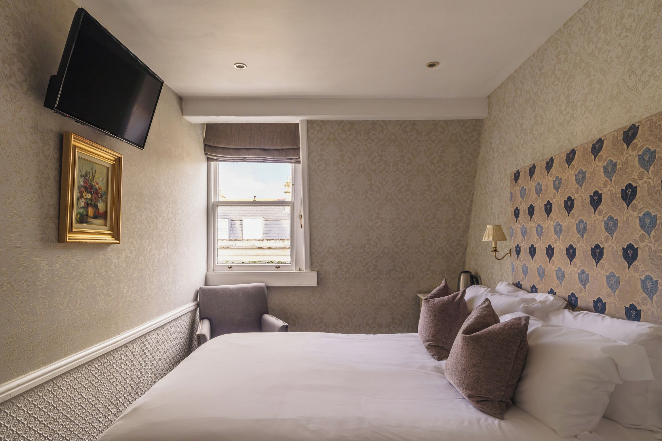 A hotel room with a large bed, a television mounted on the wall, a window with a view of the city, and patterned wallpaper.