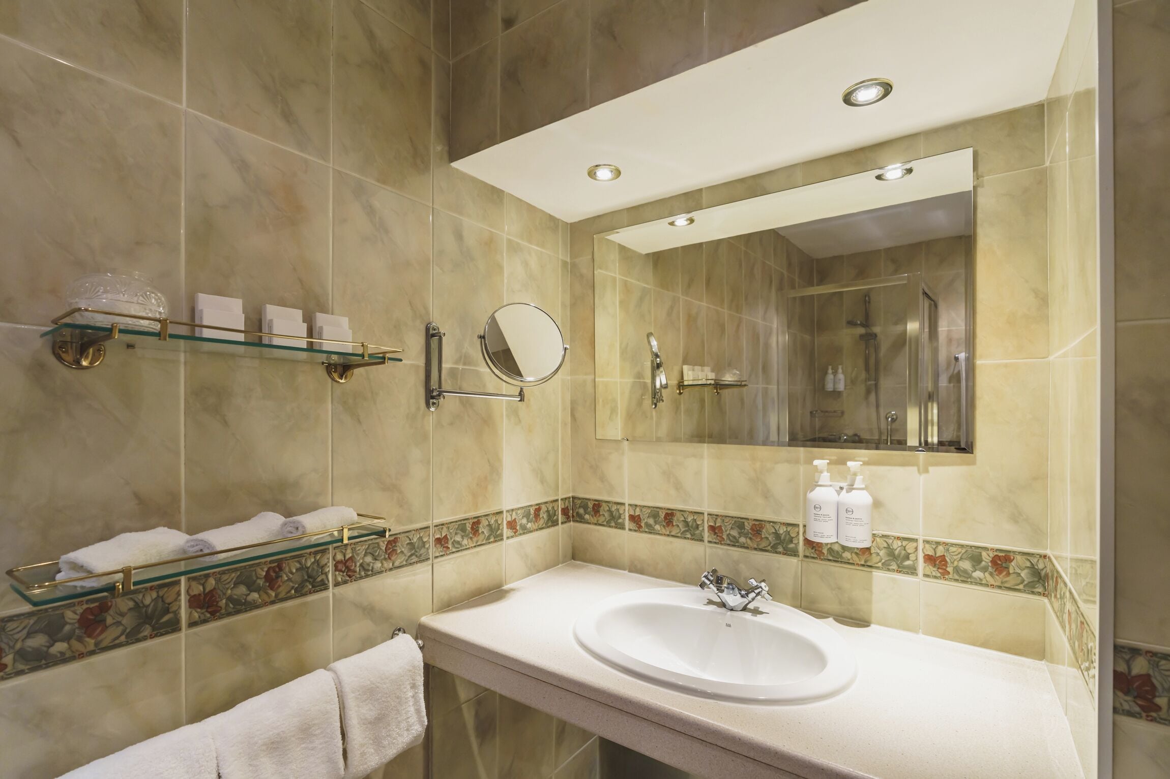 A modern bathroom with a large mirror, a glass shelf with toiletries, and a marble-tiled wall.