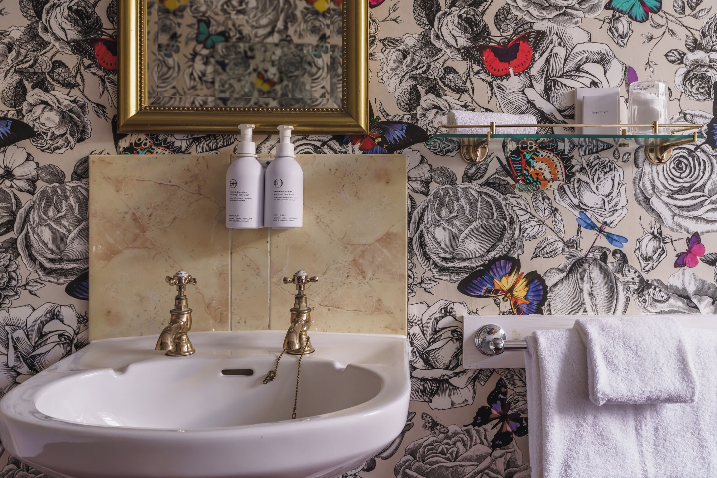 Et badeværelse med en hvid vask, to guldfarvede vandhaner, et indrammet spejl og en hylde med toiletartikler.
