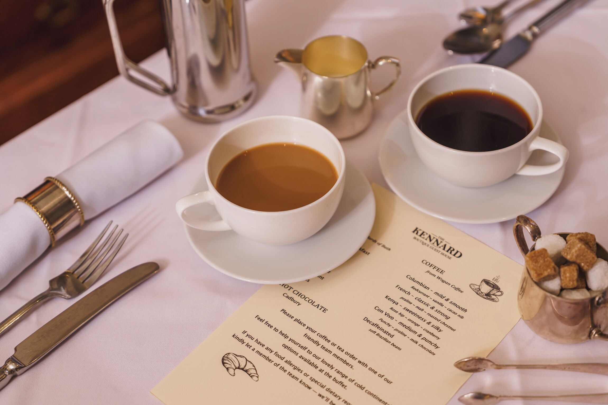 A table set with a menu, two cups of coffee, a silver pitcher, and a small bowl of sugar cubes.