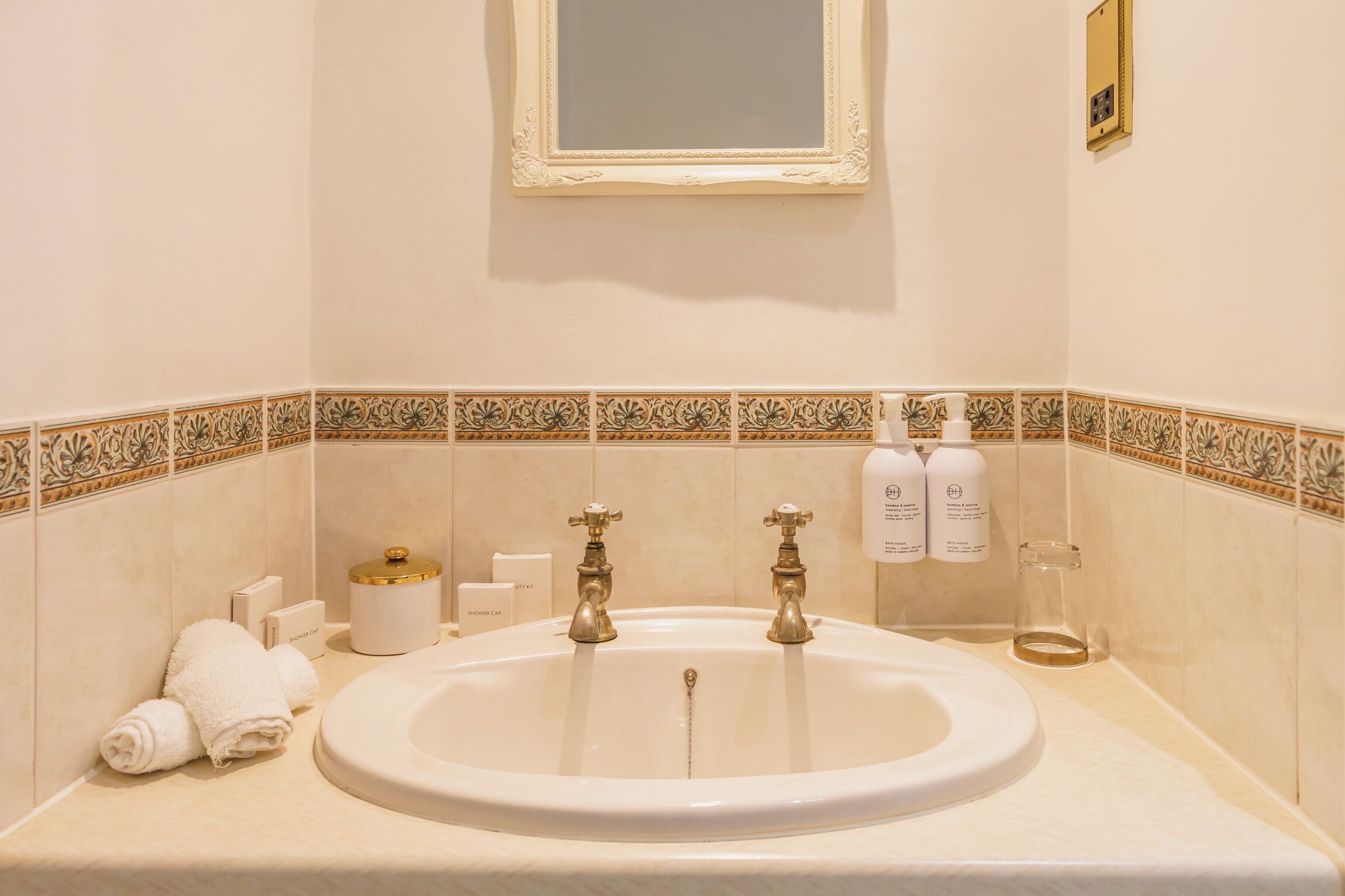 A bathroom with a white sink, two gold faucets, and a mirror above it. There are decorative tiles along the wall, a glass of water, and a bottle of shampoo.