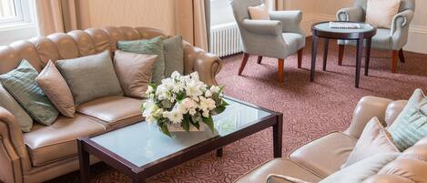 Una sala de estar bien amoblada con un sofá de cuero color beige, una mesa de centro de vidrio con un arreglo floral y dos sillones.