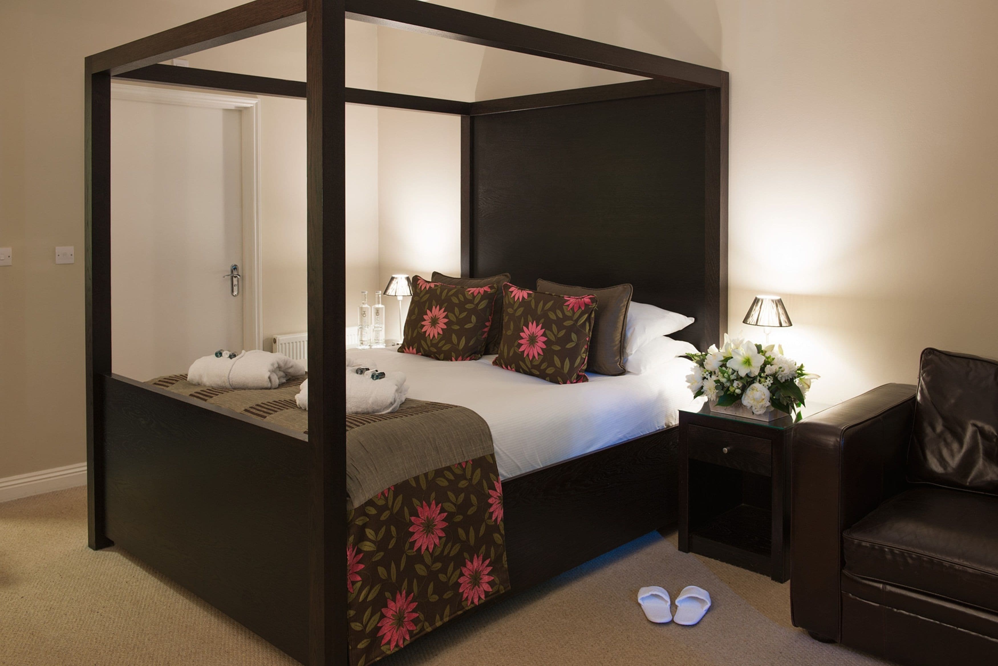 Una cama con dosel de estructura oscura, ropa de cama blanca y almohadas estampadas con motivos florales. Hay una mesita de noche con una lámpara y un jarrón de flores. Se ve una silla de cuero marrón a la derecha.