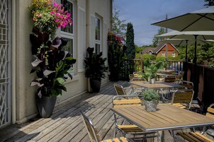 An outdoor patio area with wooden tables and chairs, potted plants, and a large umbrella.