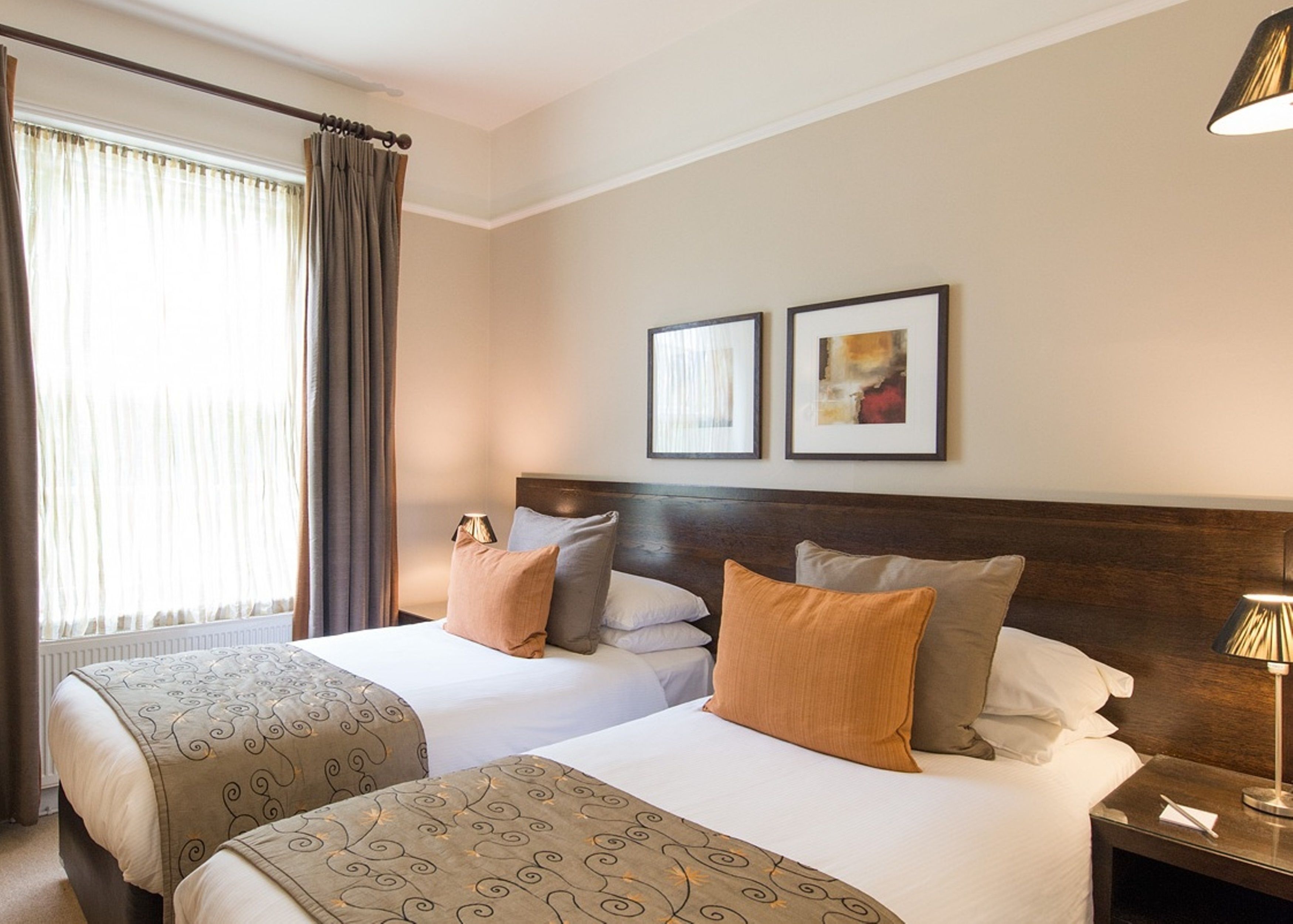 A hotel room with two beds, a large window with curtains, a wooden headboard, and two framed pictures on the wall.