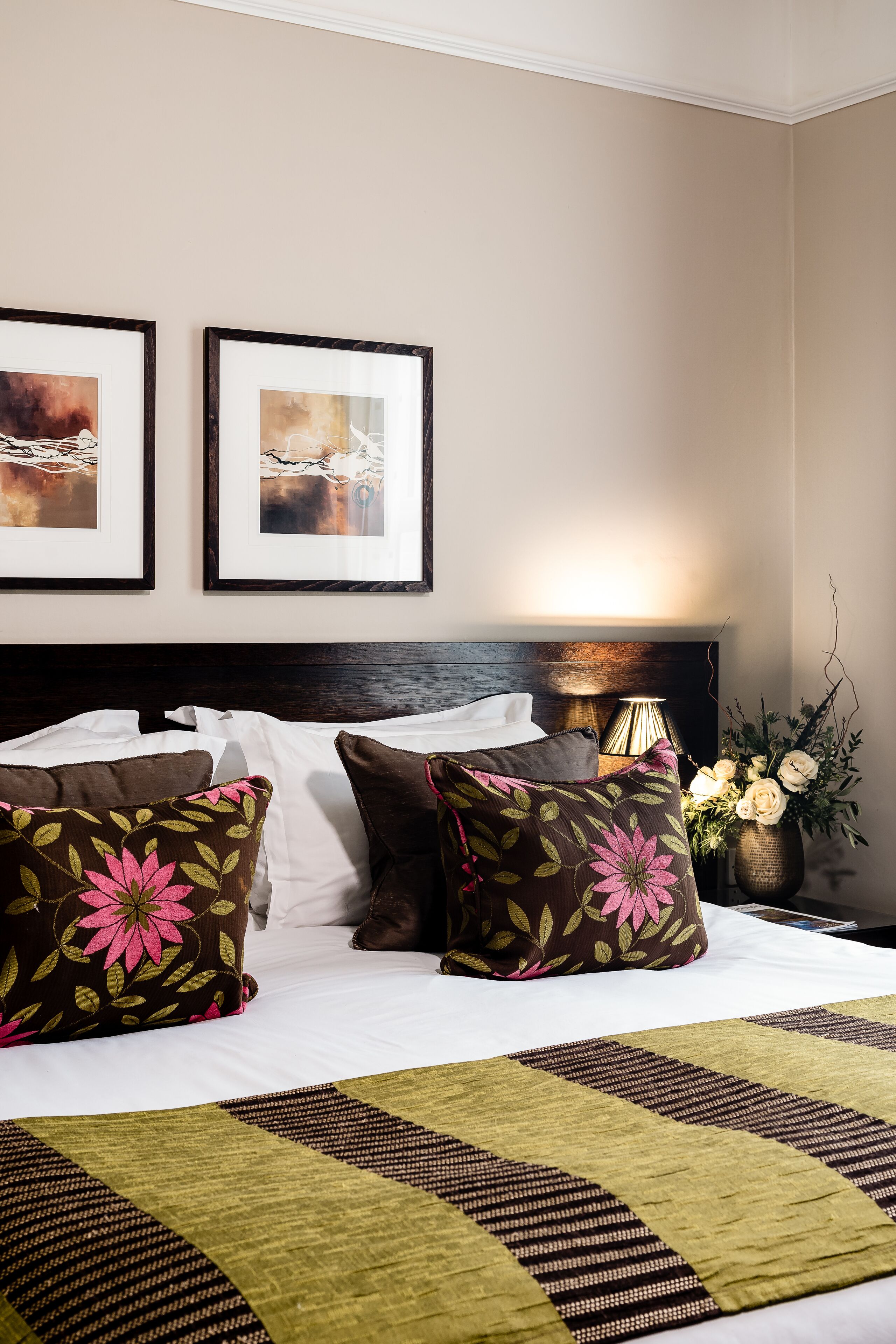 A neatly made bed with floral pillows, a bedside lamp, and a vase of flowers.