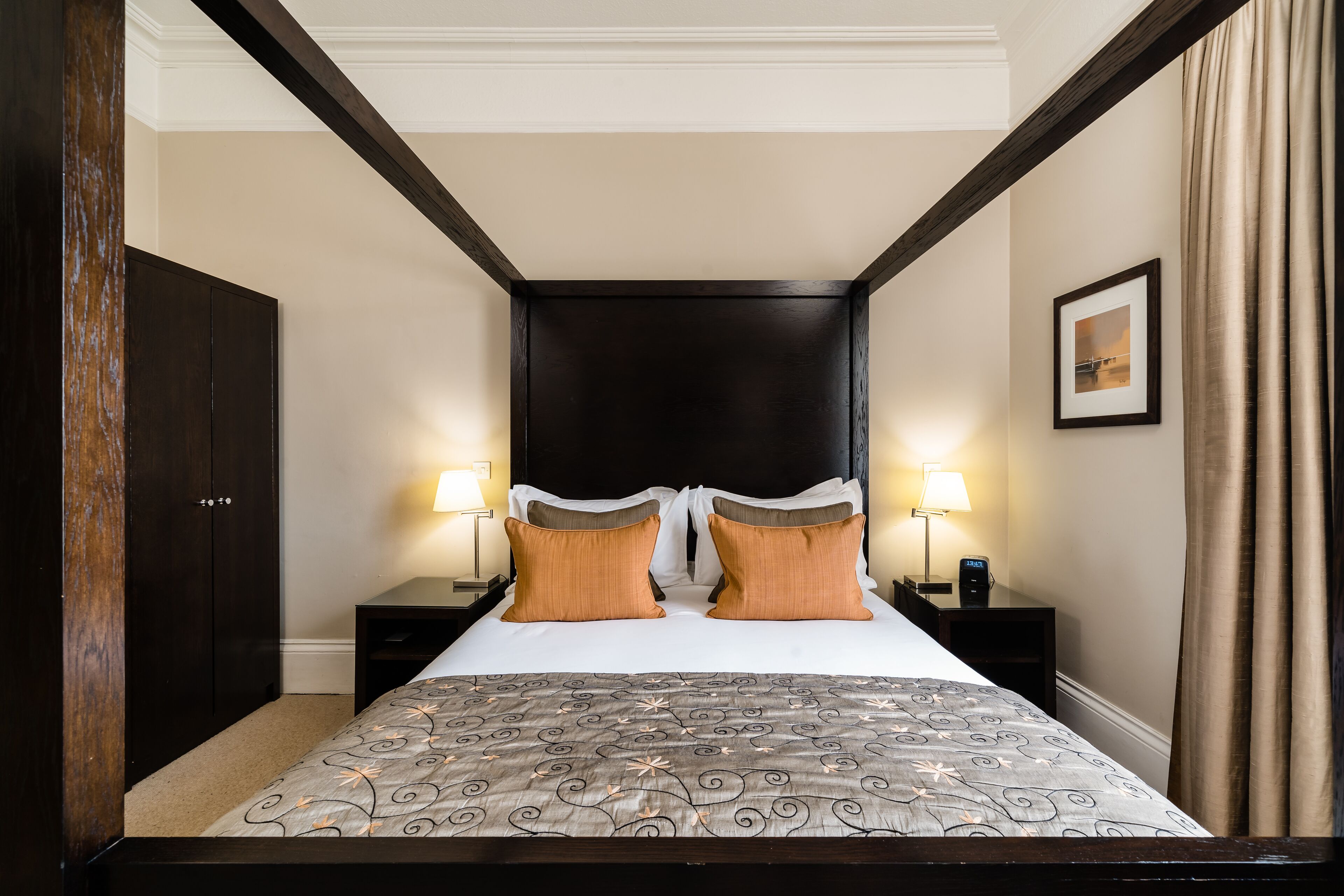 Habitación de hotel con una cama grande, cabecera de madera oscura, dos mesitas de noche con lámparas y un cuadro en la pared.