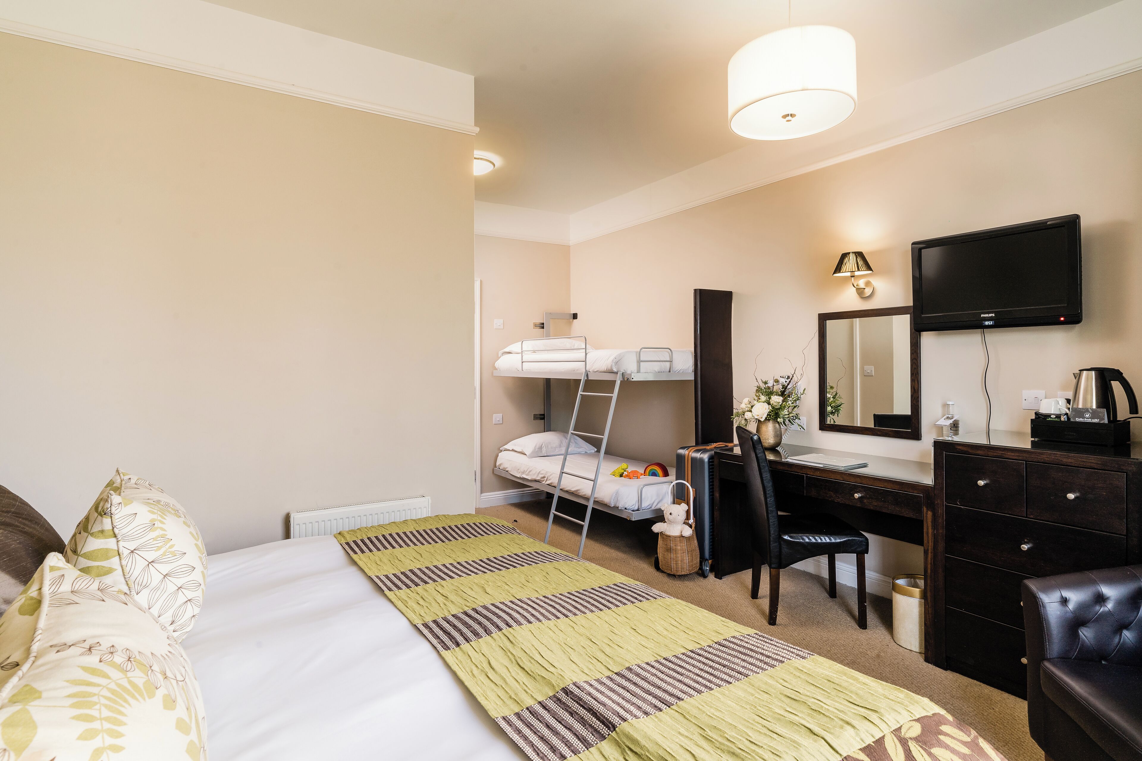 A hotel room with a bunk bed, a desk with a chair, a dresser, a television, and a lamp.