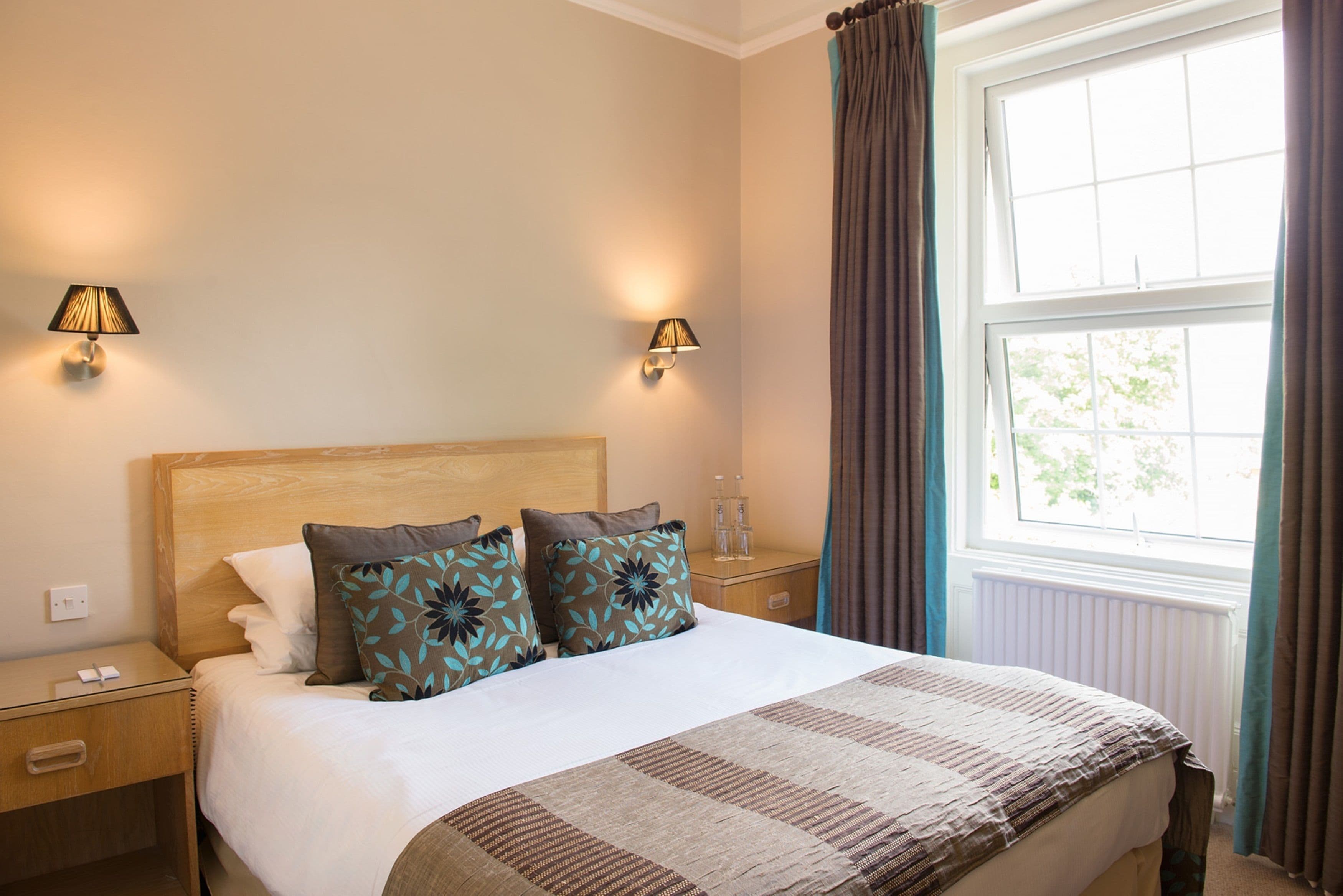 A neatly made bed with a patterned bedspread, two wall-mounted lamps, and a window with curtains.