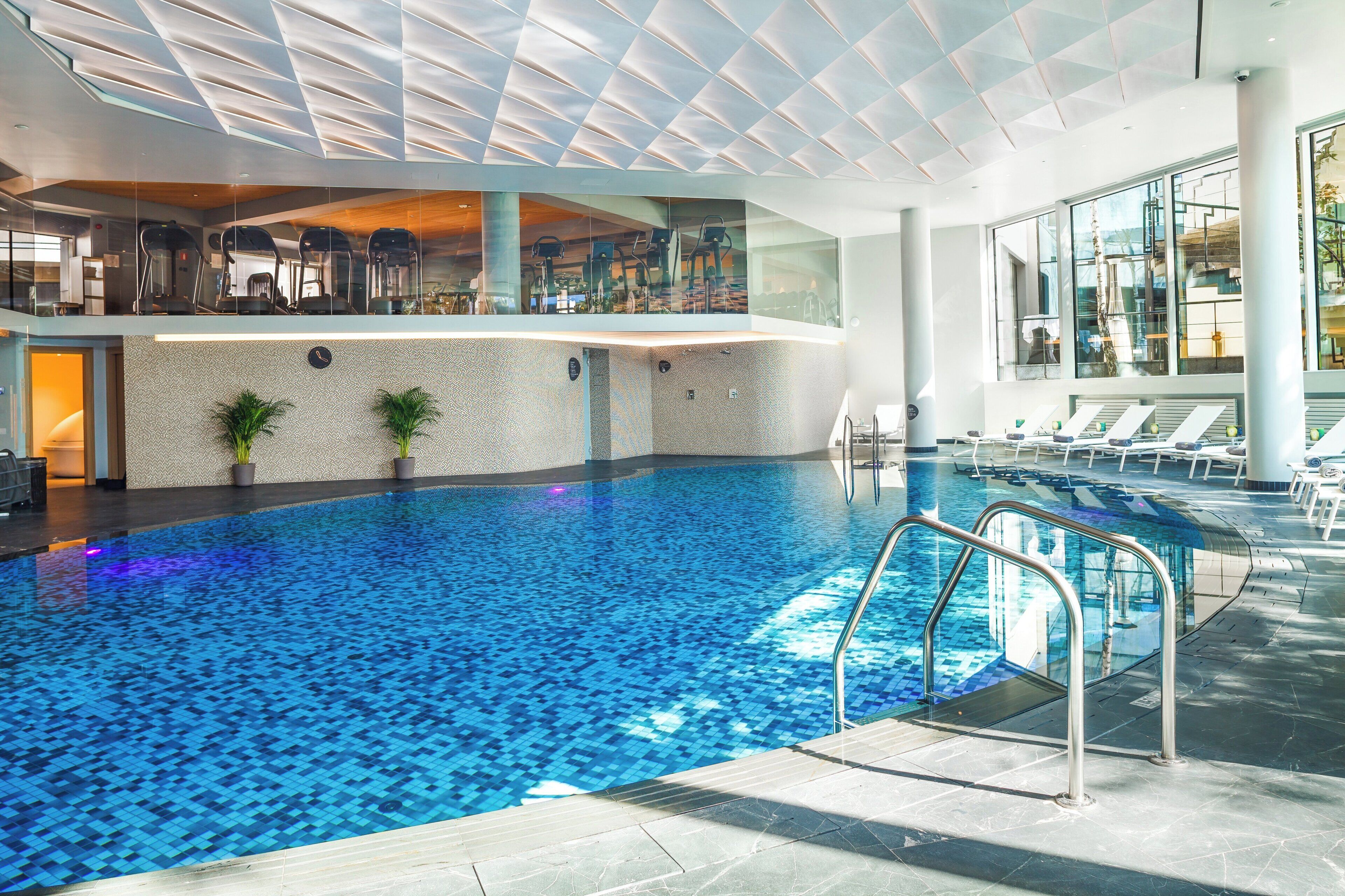 Una piscina interior con un diseño geométrico de azulejos, baranda de cristal y techo iluminado por el sol.