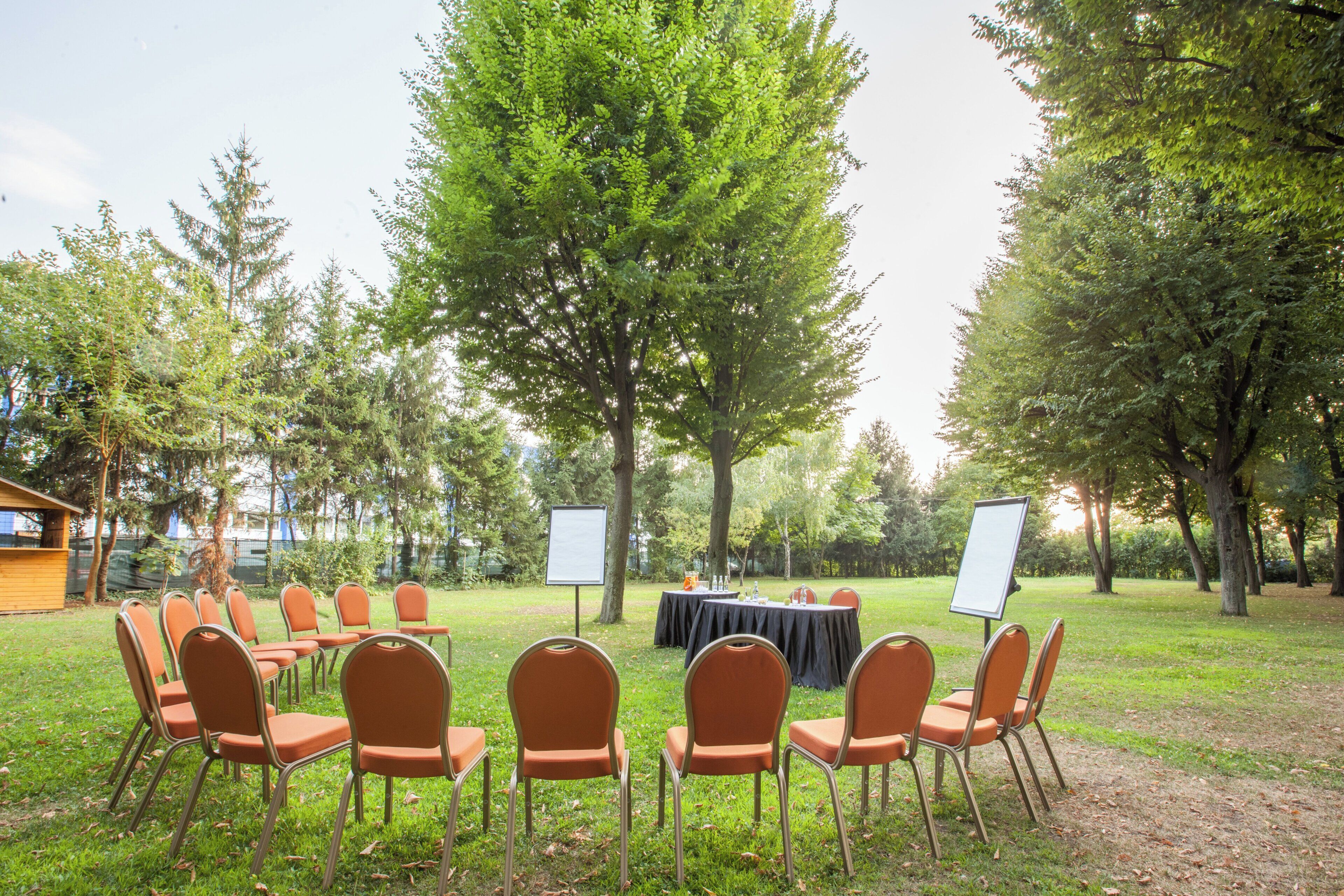 An outdoor event space with a table and chairs set up for a meeting or presentation, surrounded by trees.