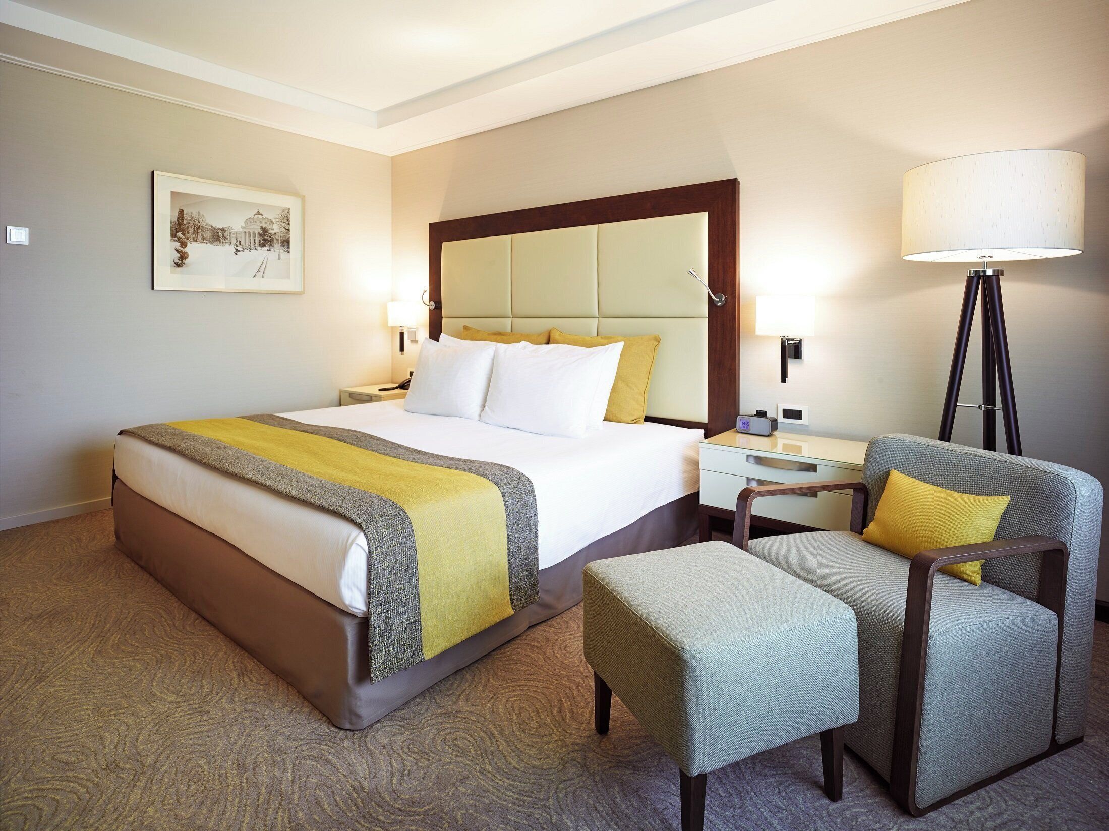 A hotel room with a bed, bedside table, lamp, chair, and a framed picture on the wall.