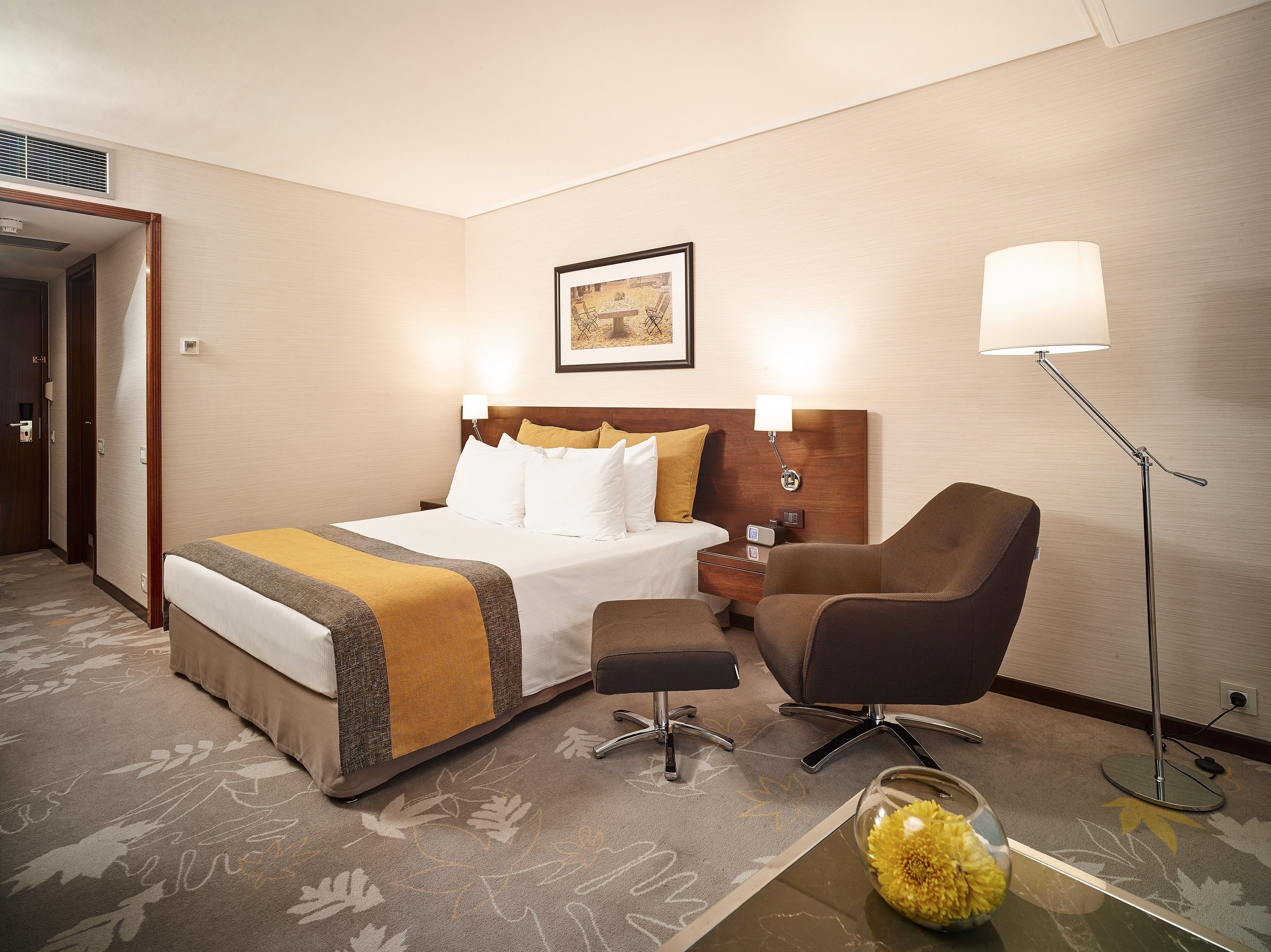 A hotel room with a bed, a desk, a chair, a floor lamp, and a vase with flowers.
