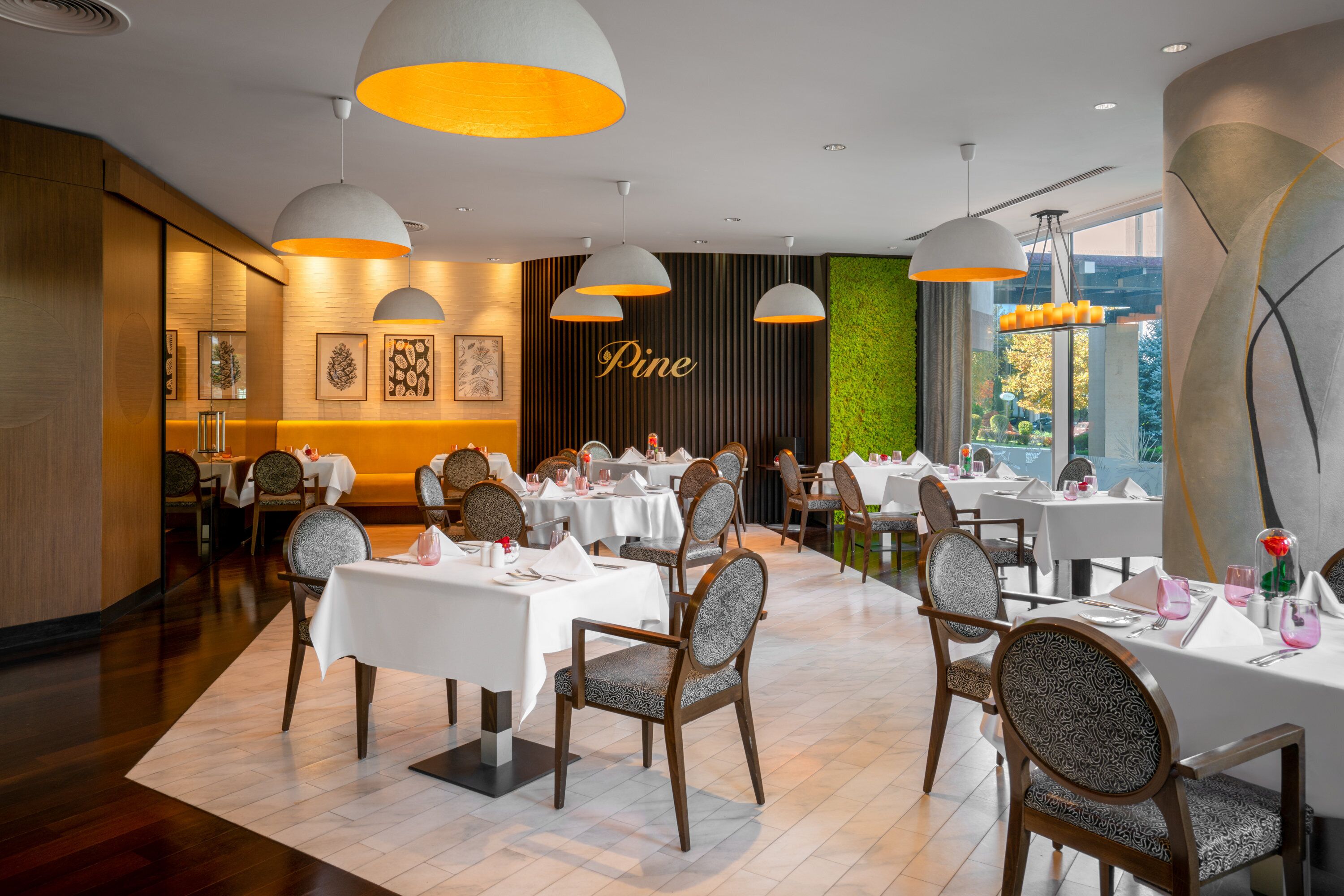 A restaurant with white tables and chairs, a green wall with the word "Pine" on it, and pendant lights.