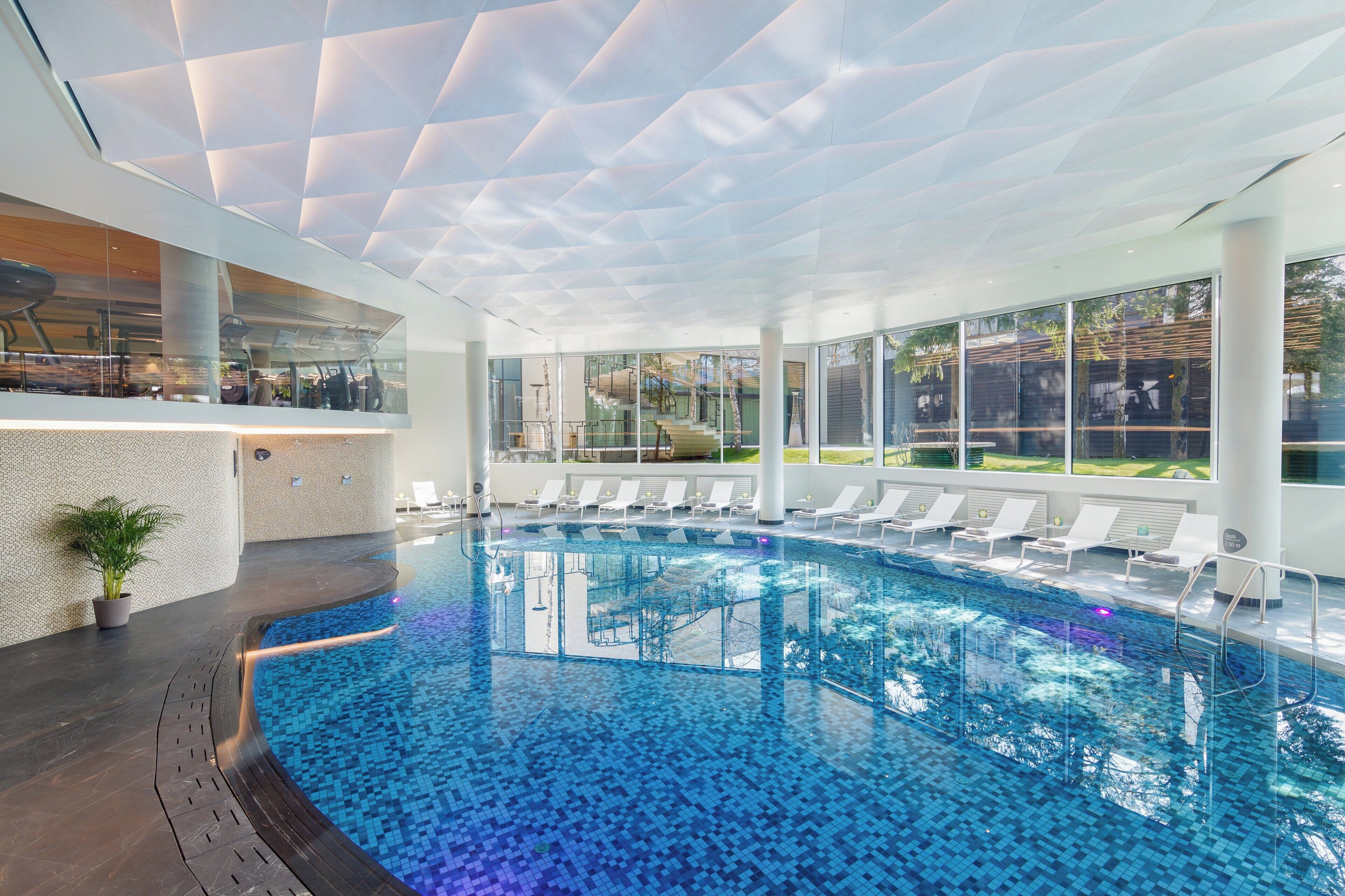 A modern indoor swimming pool with clear blue water, surrounded by lounge chairs and a glass wall offering a view of the outdoors.