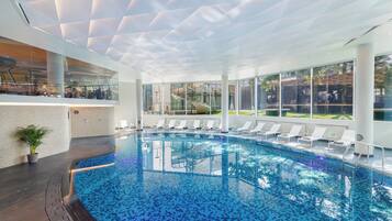 A modern indoor swimming pool with clear blue water, surrounded by lounge chairs and a glass wall offering a view of the outdoors.