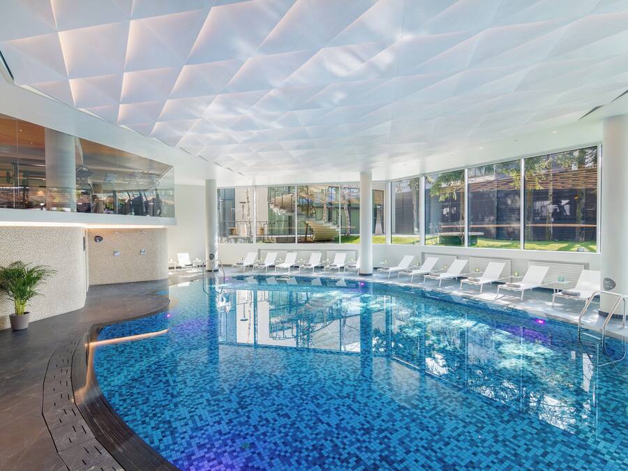 A modern indoor swimming pool with clear blue water, surrounded by lounge chairs and a glass wall offering a view of the outdoors.