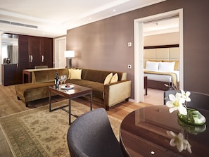 A modern hotel room with a brown sofa, a dining table, a small coffee table, and a separate seating area with a chair.