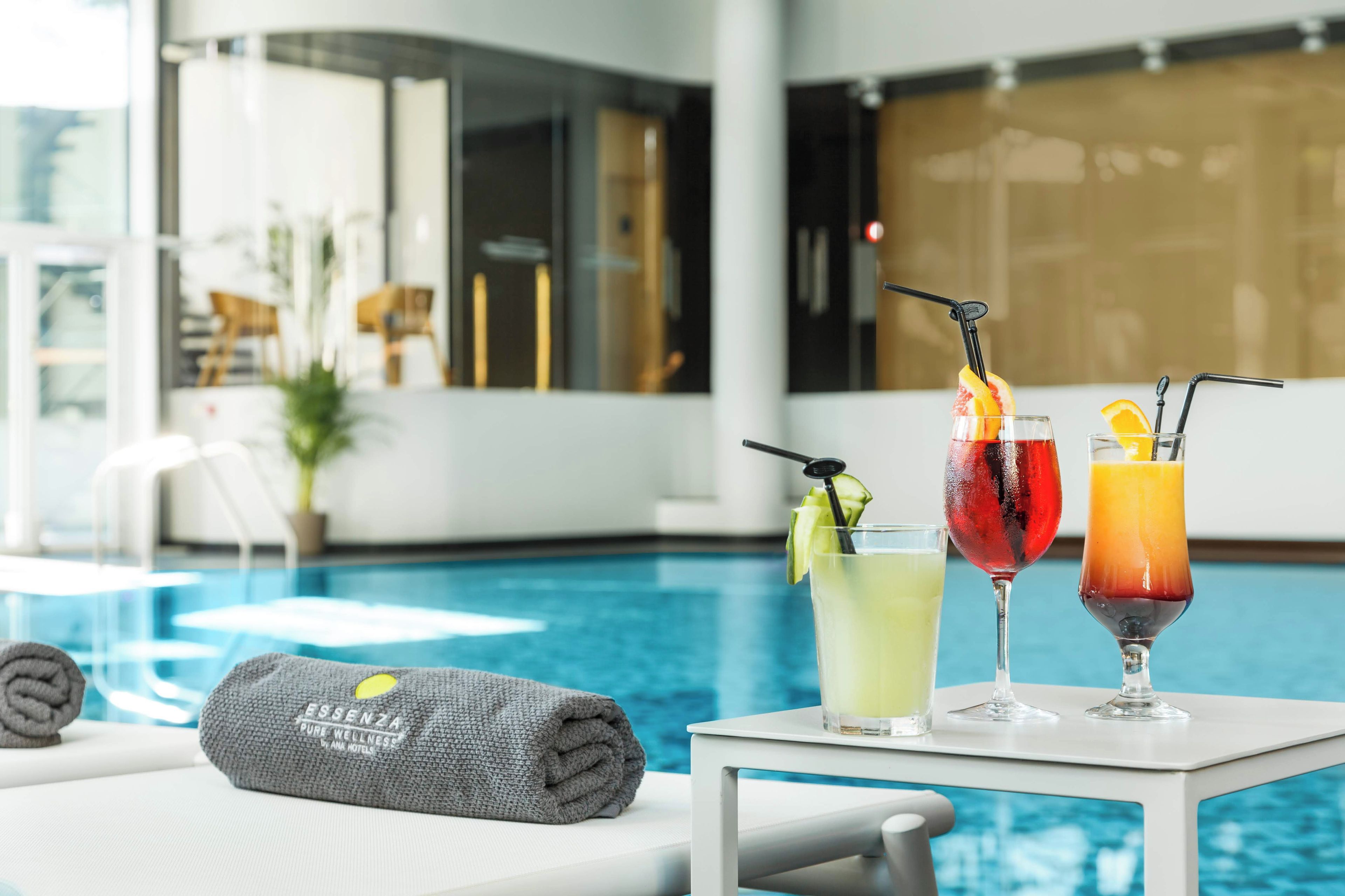 A poolside table with three drinks, a towel, and a view of the pool area.