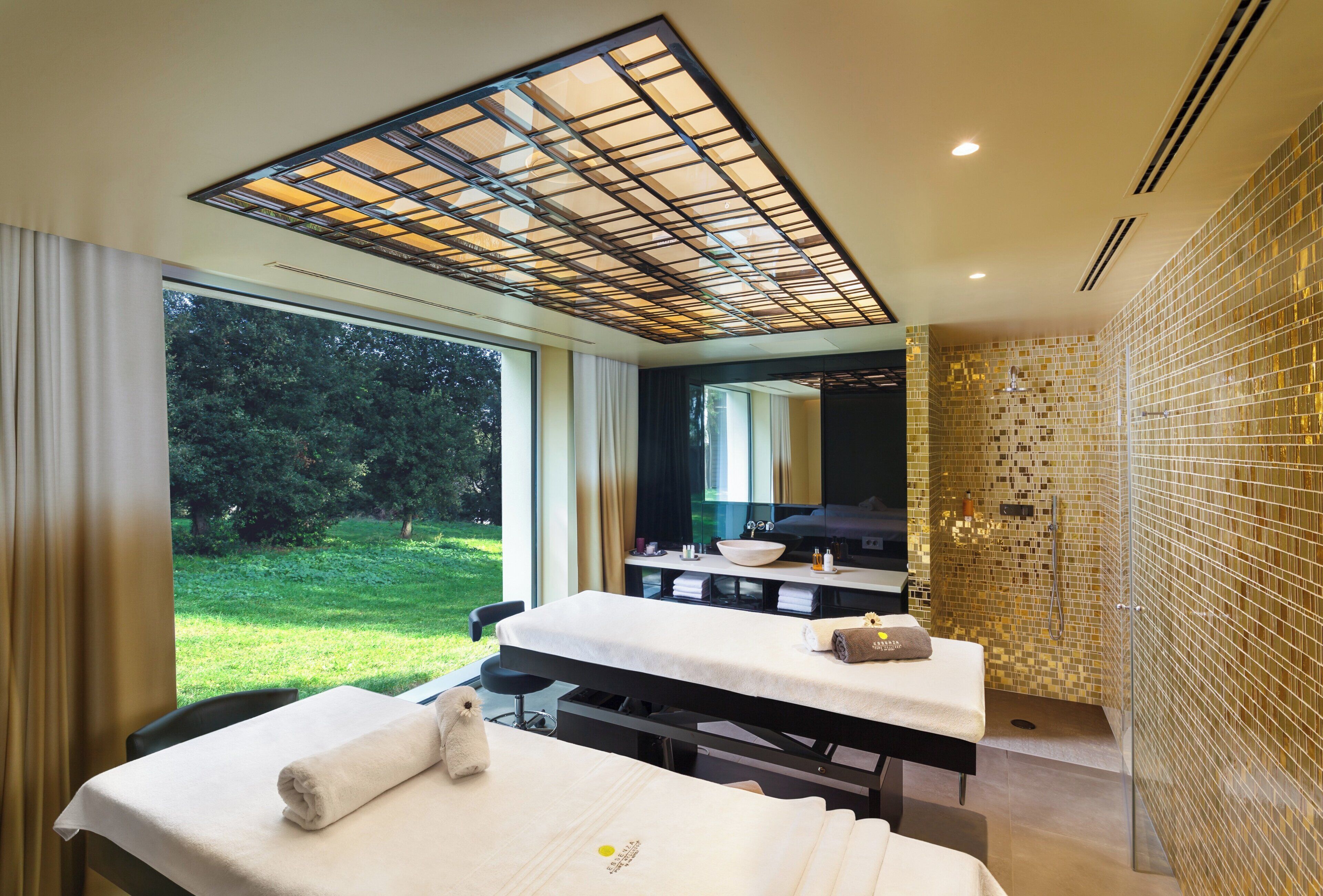 A modern spa room with a large window, a massage table, a sink, and a shower area.