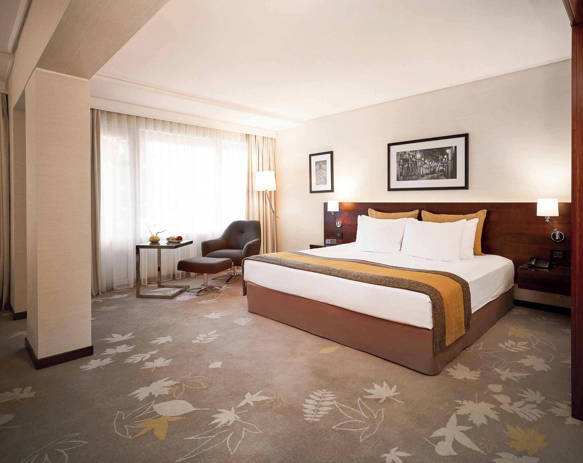 Quarto de hotel com uma cama grande, uma cadeira, uma mesinha e uma luminária.