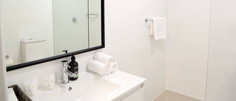 Un baño moderno con mampara de vidrio, un lavamanos blanco con artículos de aseo y un perchero para toallas.