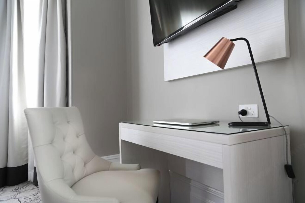 Una habitación moderna con un escritorio blanco, una silla acolchada, un televisor de pantalla plana en la pared y una lámpara de escritorio.