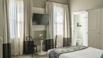 A hotel room with a bed, a TV mounted on the wall, a small table with chairs, and a bathroom visible through an open door.