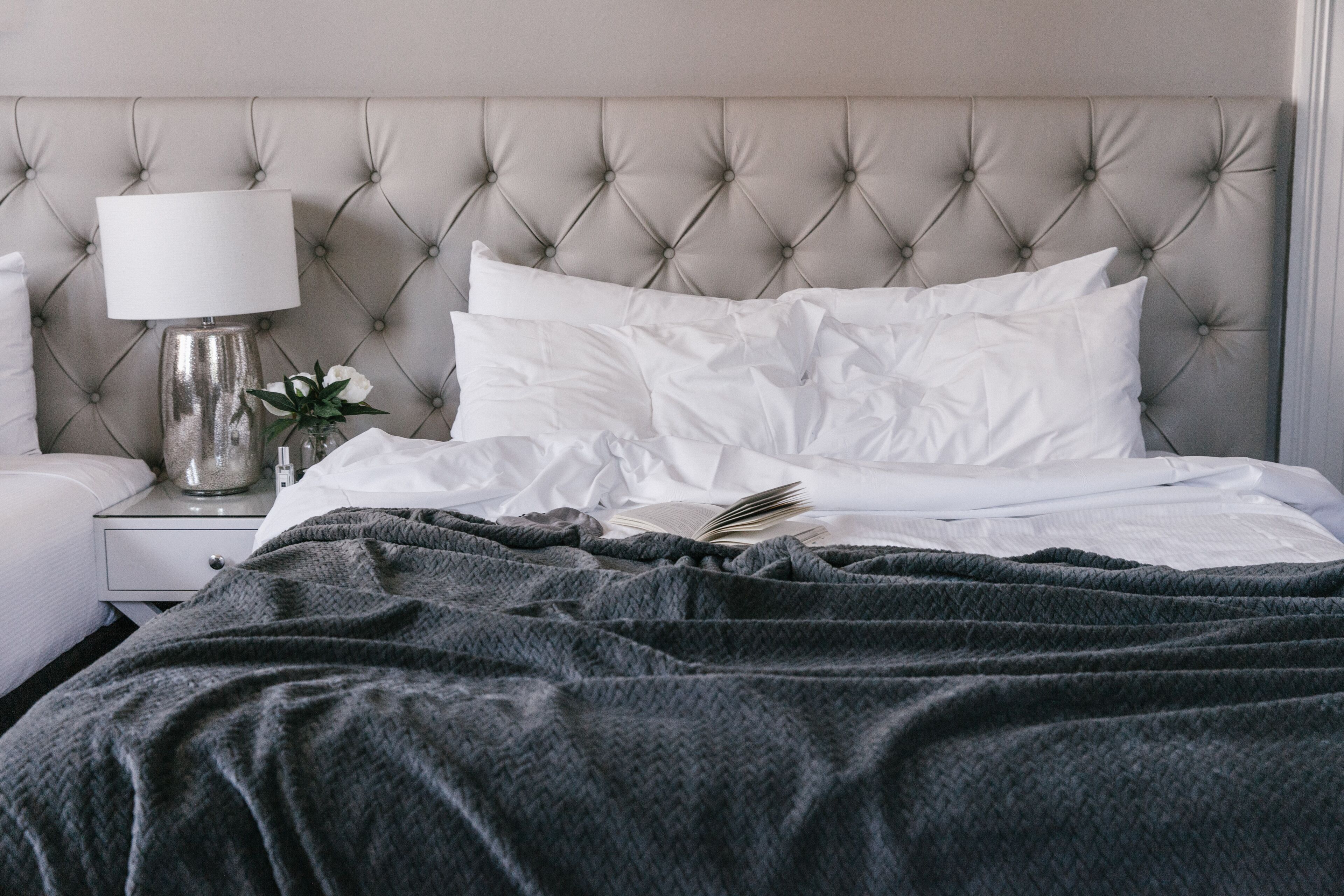 Un letto rifatto con lenzuola bianche e una coperta grigia, una lampada da comodino e un piccolo vaso con fiori su un tavolino.