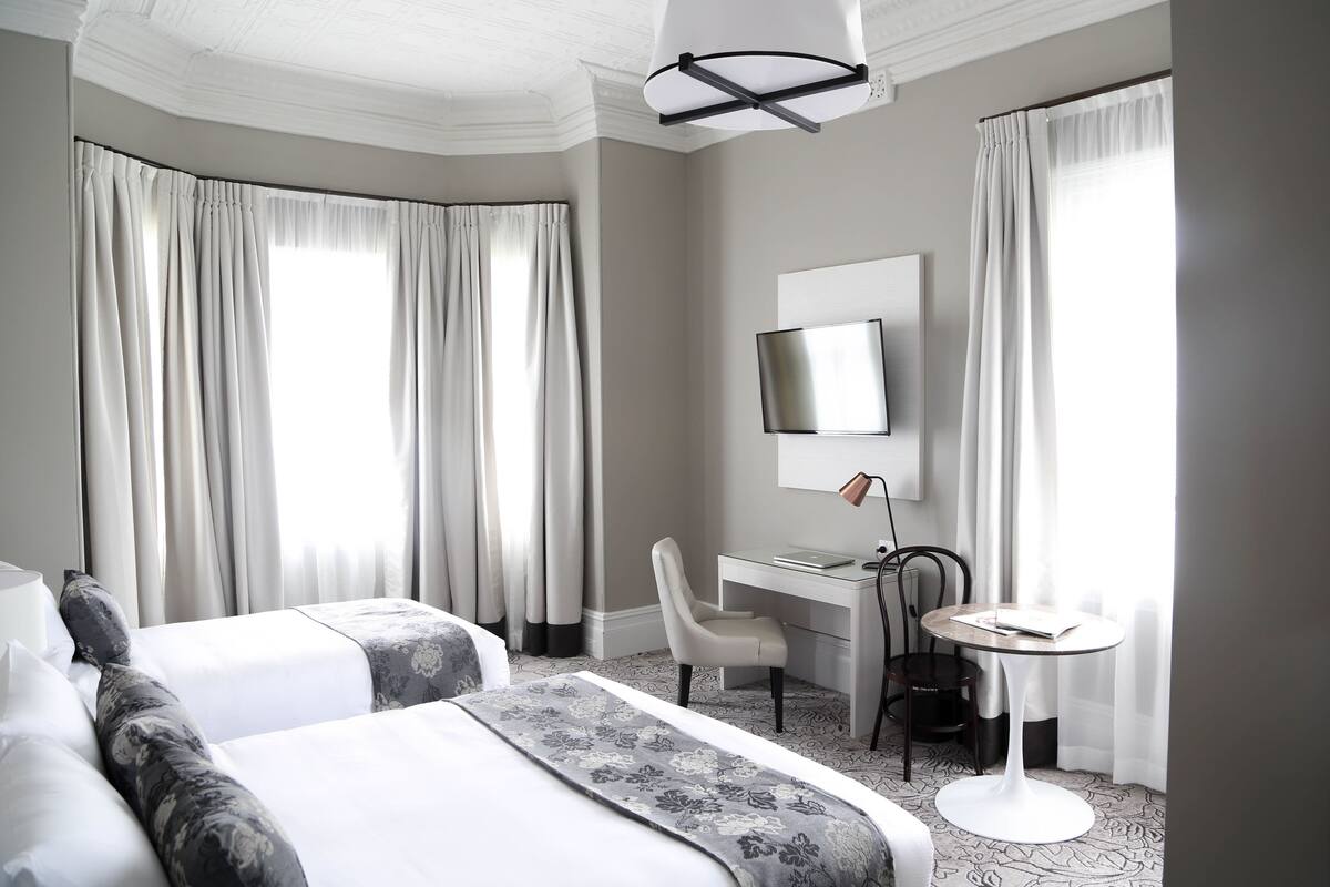 A hotel room with two beds, a desk, a chair, a small table, a TV, and large windows with curtains.