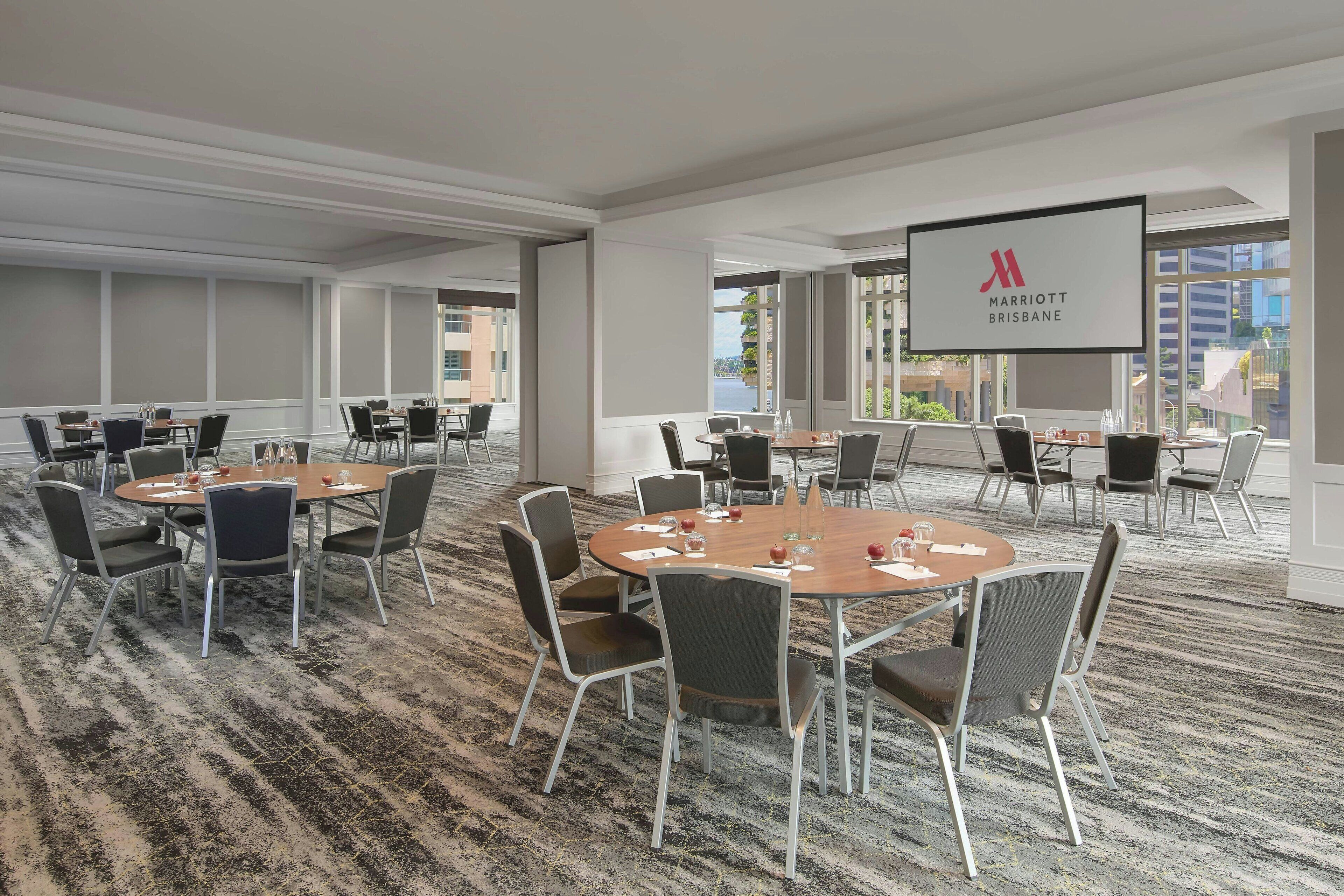Une salle de conférence dotée de plusieurs tables et chaises, de grandes fenêtres et d’un écran affichant le logo du Marriott Brisbane.