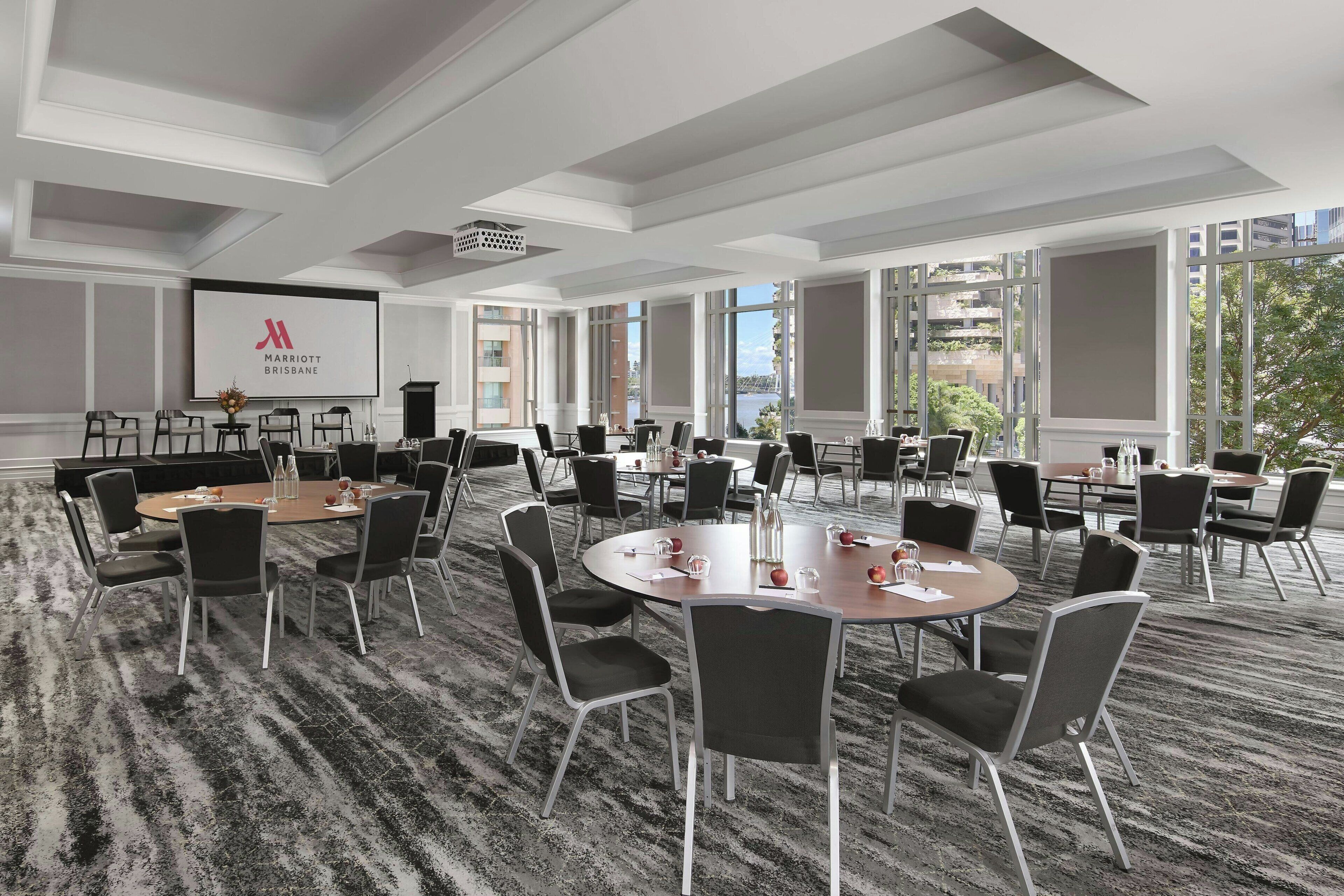 Une grande salle de conférence équipée de nombreuses tables et chaises, un écran de projection affichant le logo du Marriott Brisbane, et de grandes fenêtres offrant une vue sur la ville.