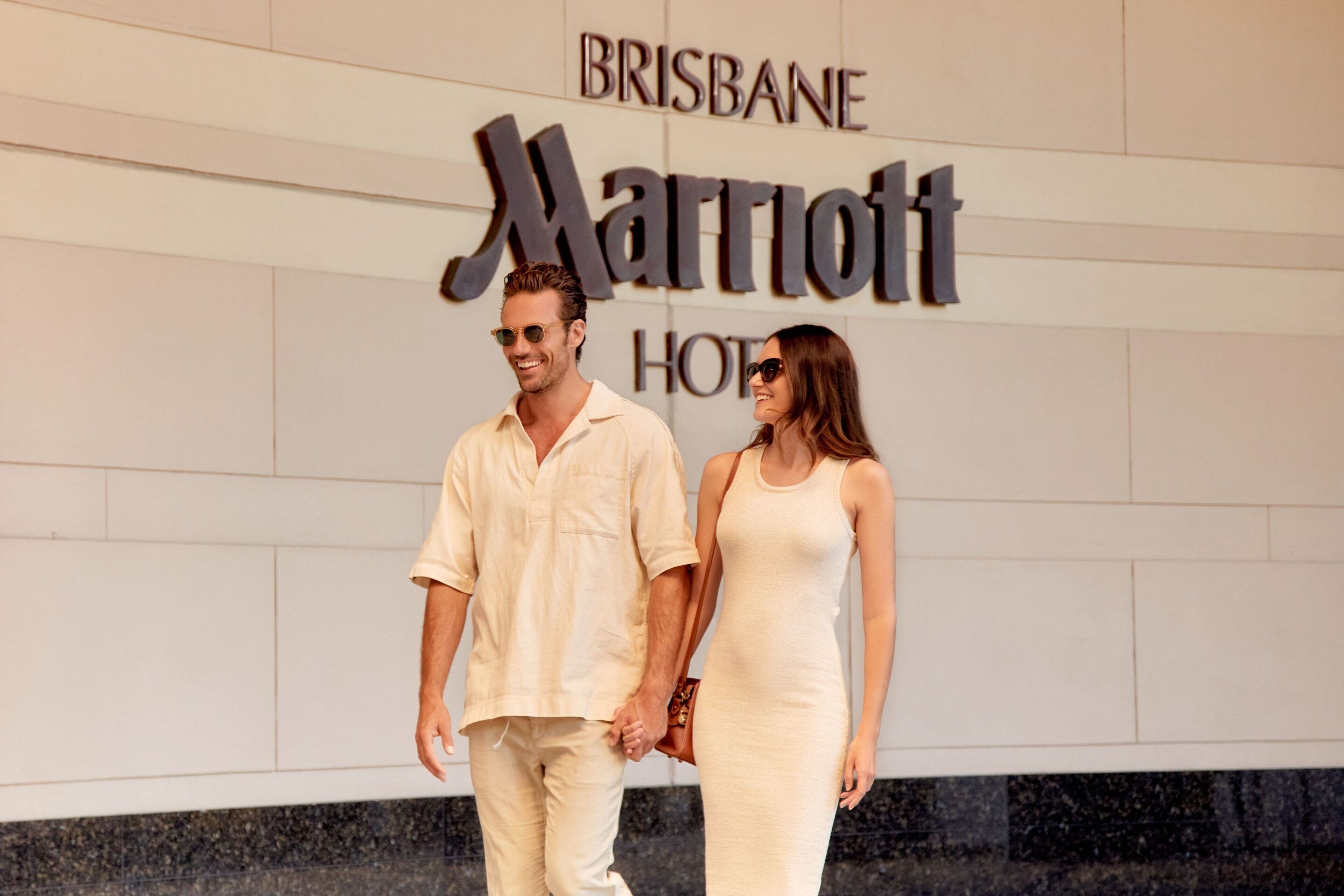 A couple walking in front of a Marriott hotel sign with the words "BRISBANE HOTEL" visible.