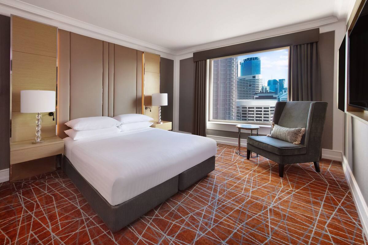Ein Hotelzimmer mit einem großen Bett, einem Sessel, einem kleinen Tisch und Blick auf die Stadt.