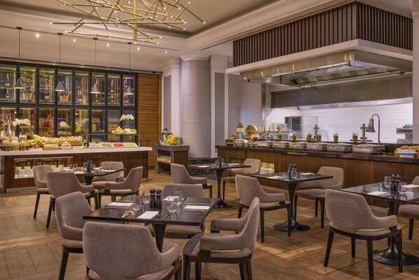 A modern restaurant with a buffet area, multiple dining tables, and a visible kitchen area.