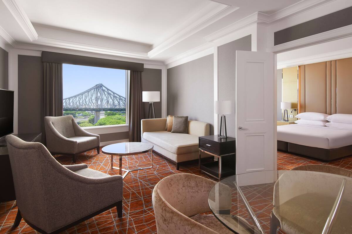 Ein Hotelzimmer mit Sofa, Sesseln, einem kleinen Tisch, einem Bett und Blick auf eine Brücke.
