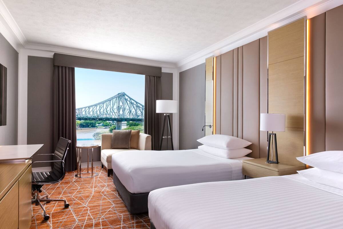 Ein Hotelzimmer mit zwei Betten, einem Schreibtisch, einem Stuhl und Blick auf eine Brücke.