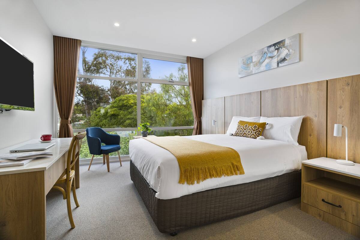 A modern hotel room with a large bed, a desk, a chair, and a TV.