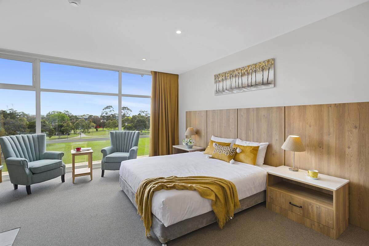 A modern hotel room with a large bed, two armchairs, a small table, and a view of a park.