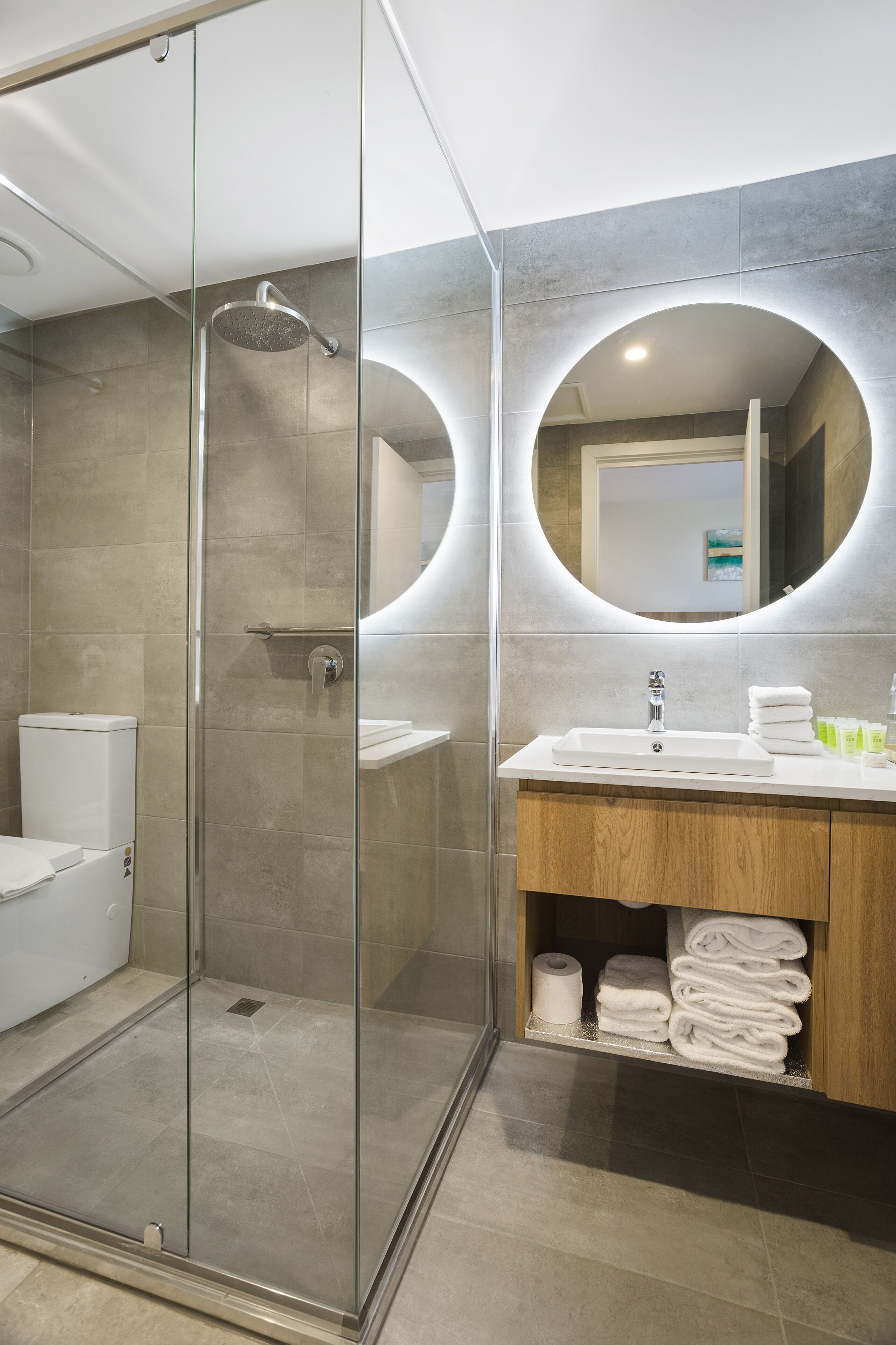 A modern bathroom with a glass-enclosed shower, a wooden vanity with a sink, and a mirror with a circular light.