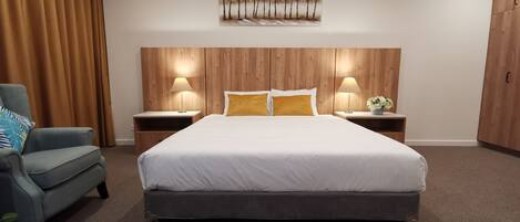 A modern hotel room with a large bed, two bedside tables with lamps, a wooden headboard, a blue armchair, and a wall-mounted artwork.