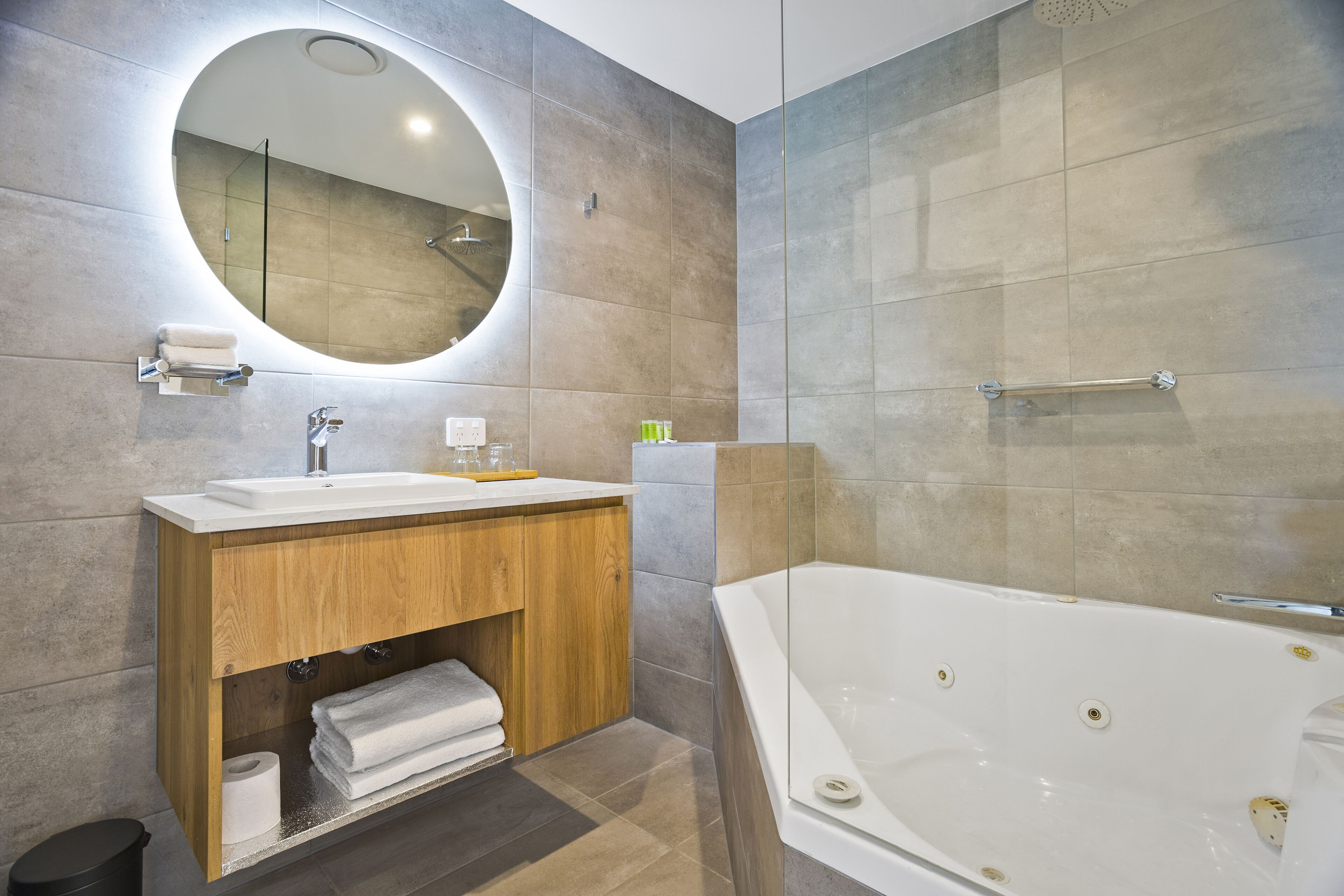 A modern bathroom with a large oval mirror, a wooden vanity, a white bathtub, and a shower area.