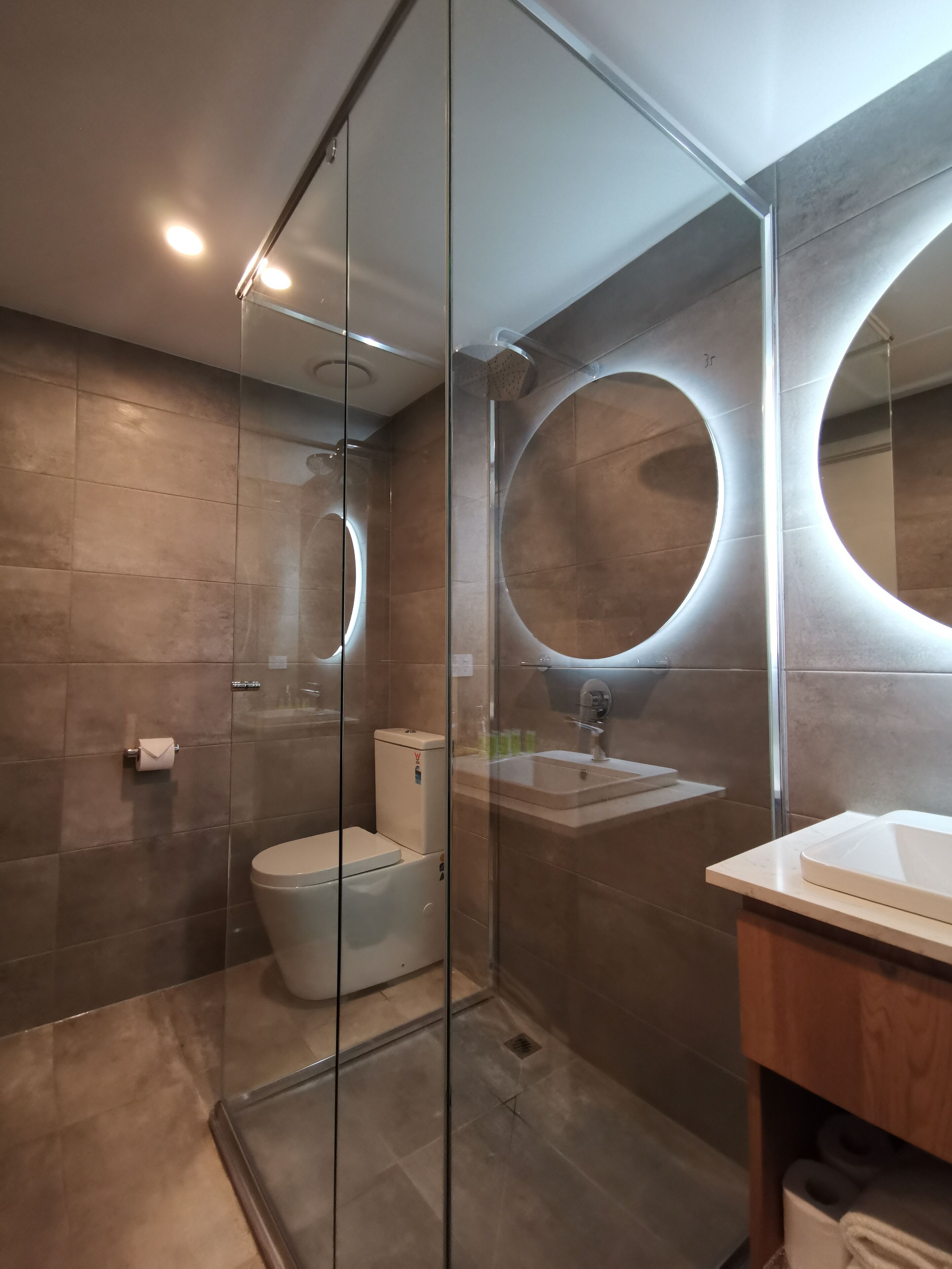 A modern bathroom with a glass shower enclosure, a toilet, a sink, and a large mirror with circular lighting.