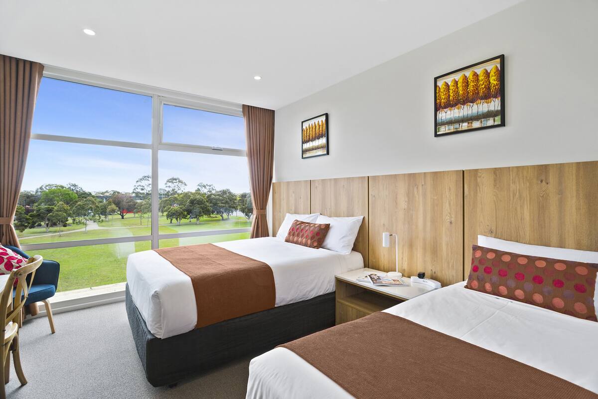 A hotel room with two beds, a wooden headboard, a large window with a view of a golf course, and a framed picture on the wall.