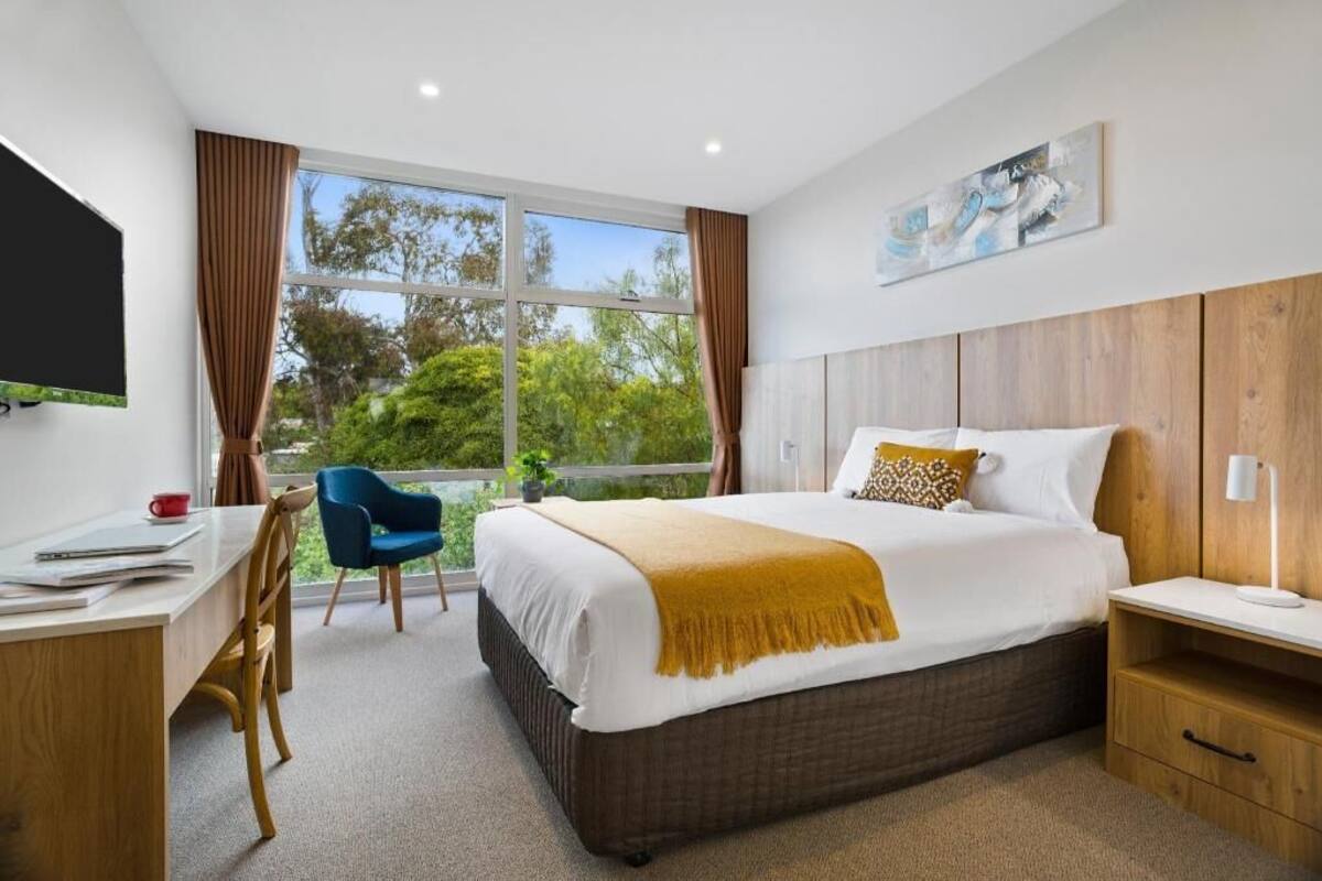 A modern hotel room with a large bed, a desk, a chair, a TV, and a window with curtains.