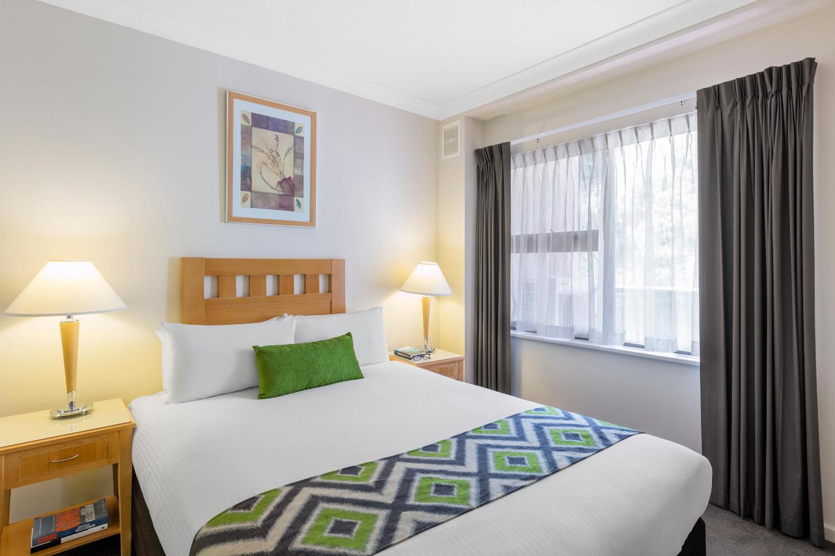 Habitación de hotel con cama, dos lámparas de noche, mesita de noche, ventana con cortinas e imagen enmarcada en la pared.