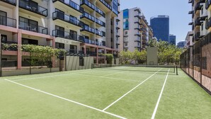 A tennis court surrounded by multi-story buildings with balconies and a clear sky. - Nesuto Mounts Bay (Perth)