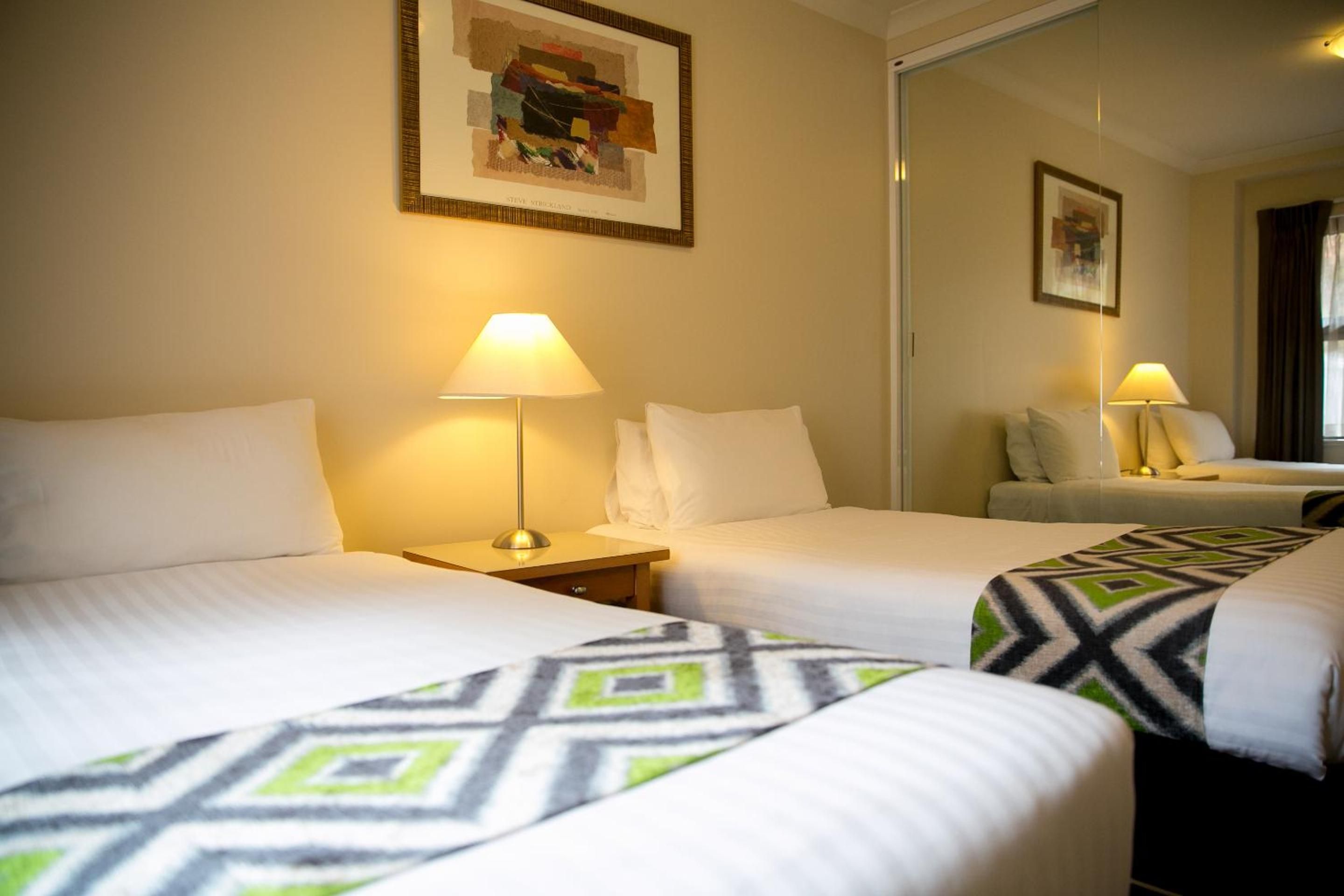 Een hotelkamer met twee bedden, elk met een kussen en een lamp. Aan de muur hangt een schilderij en een spiegel reflecteert de kamer.