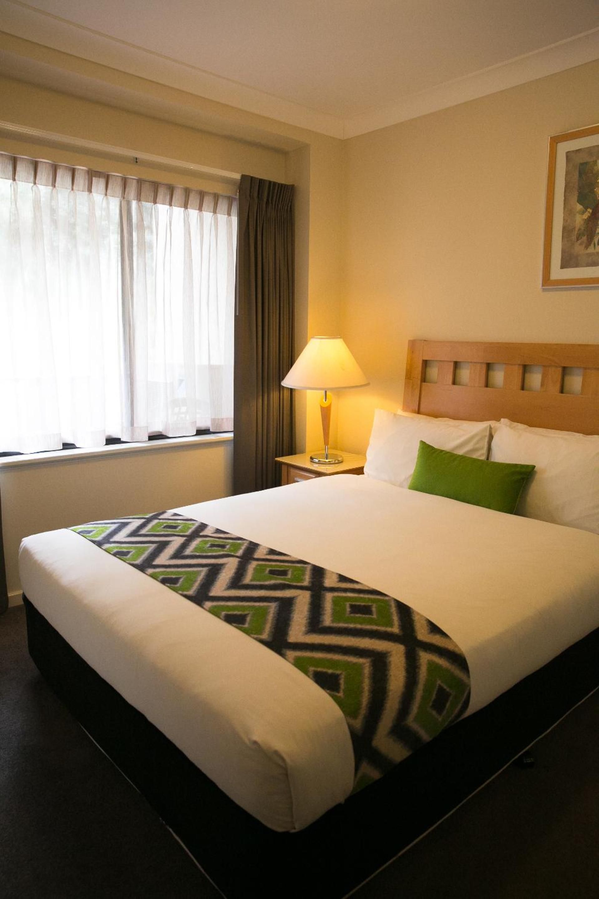 Habitación de hotel con una cama grande, una mesita de noche con lámpara, una ventana con cortinas y un cuadro en la pared.