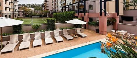 A swimming pool with sun loungers and umbrellas, surrounded by apartment buildings.